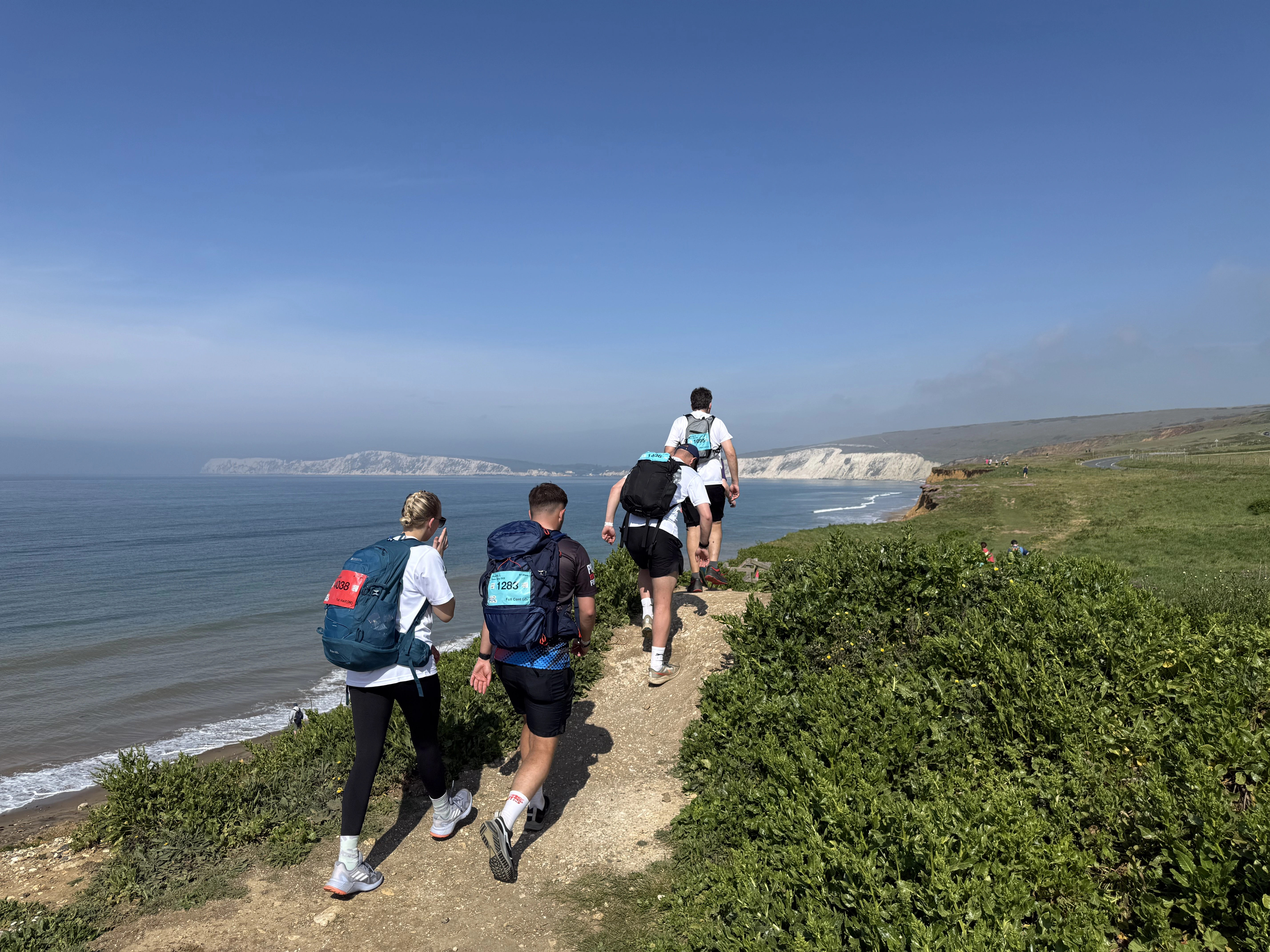 Four people walking along the Isle of Wight's coastline. They are wearing sports clothing and rucksacks, you can see white cliffs and the seaside in the background.