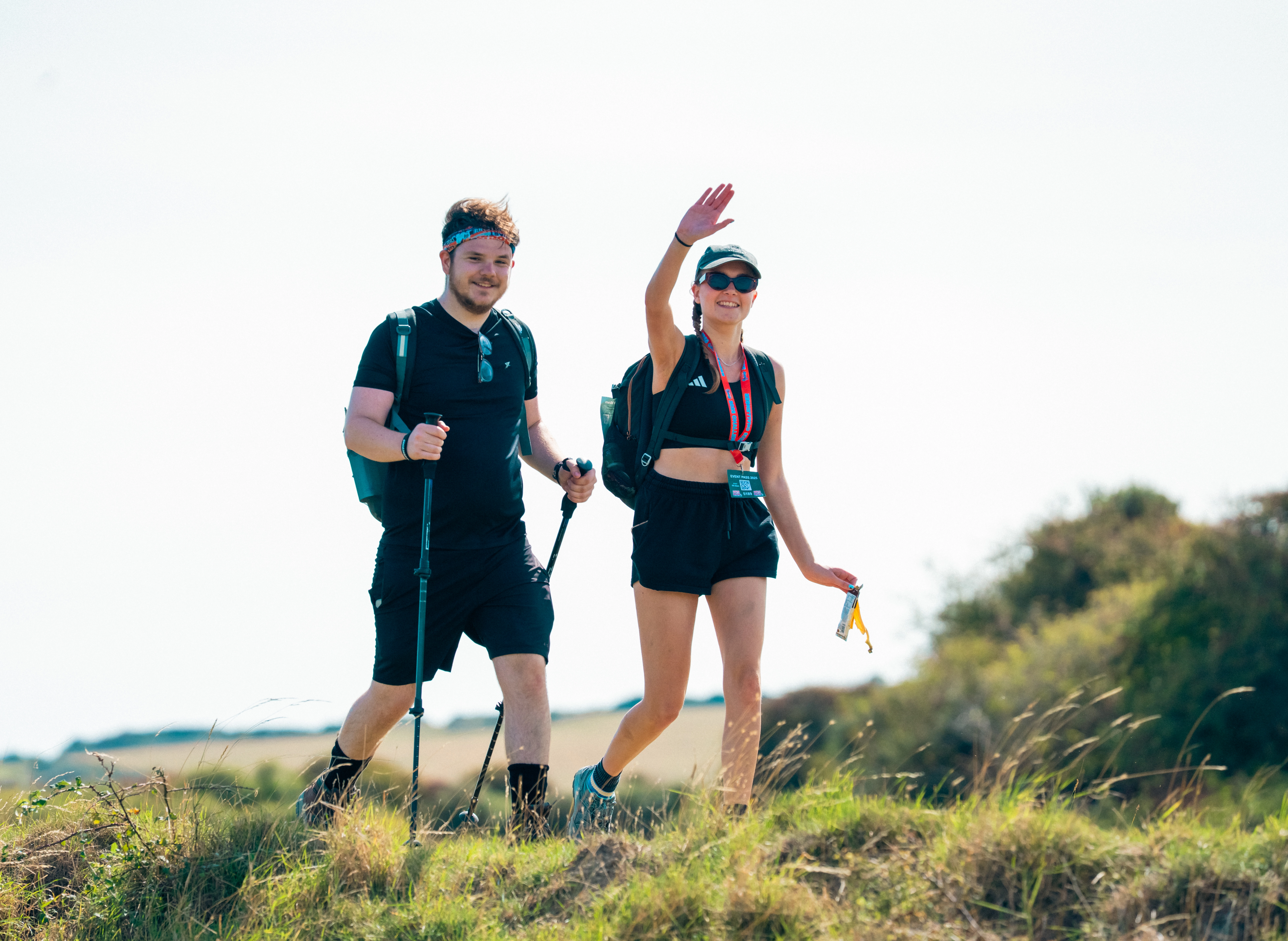 Two Ultra Challenge participants walking along in the countryside waving and smiling. One is using walking poles and the other is wearing a cap and sunglasses.
