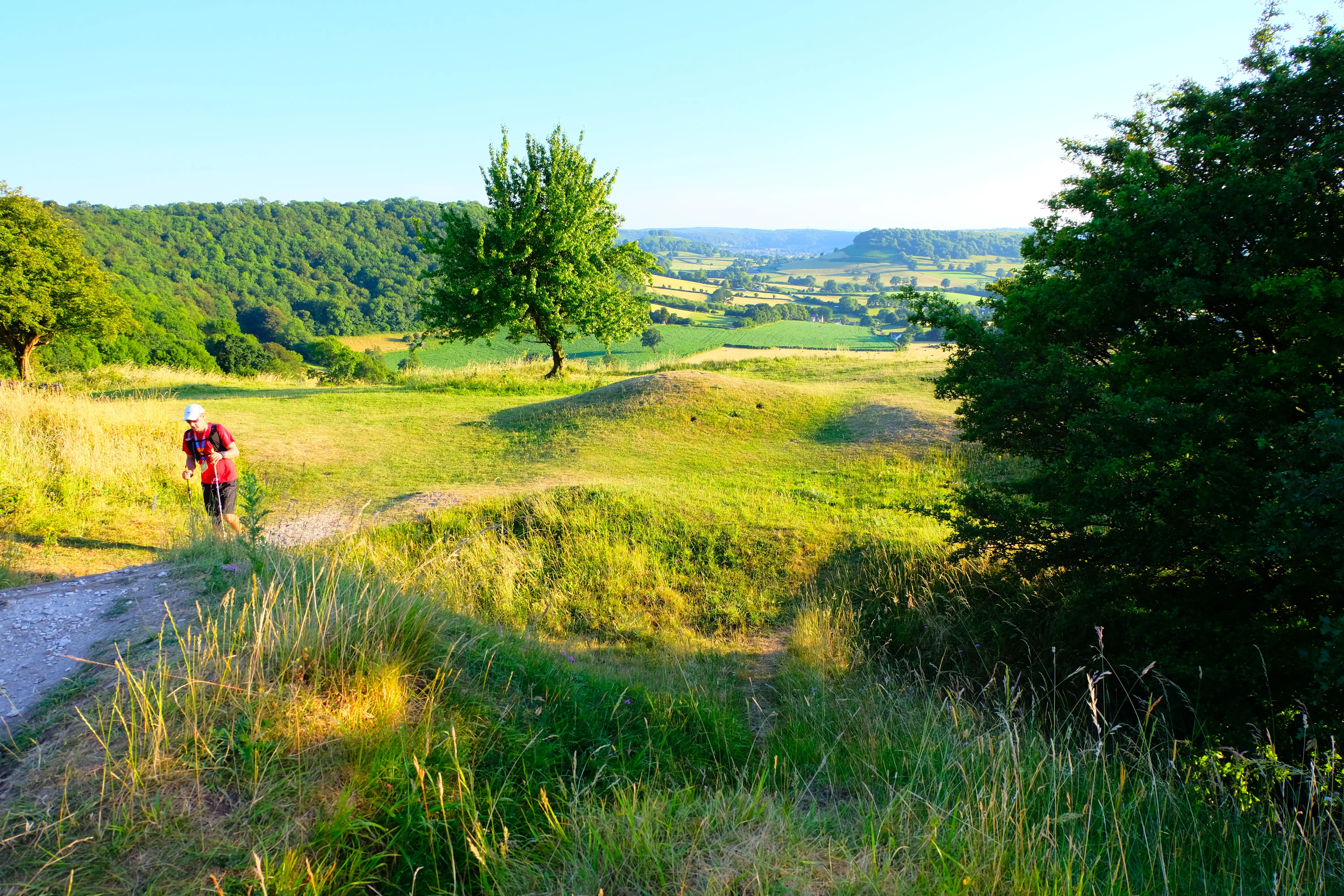 North Downs 50 Ultra Challenge participant walking through rolling fields. Large trees and hilly fields can be seen in the foreground, with trees and fields can be seen in the background.