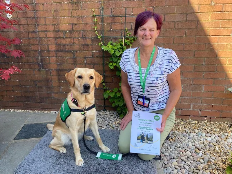 Woman in garden holding certificate and kneeling next to sitting yellow labrador