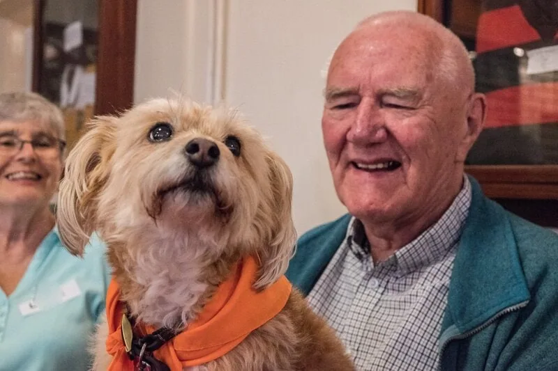 John sitting with dog Dotty on his lap. Smiling with woman sitting next to him. Dotty is wearing an orange bandana.