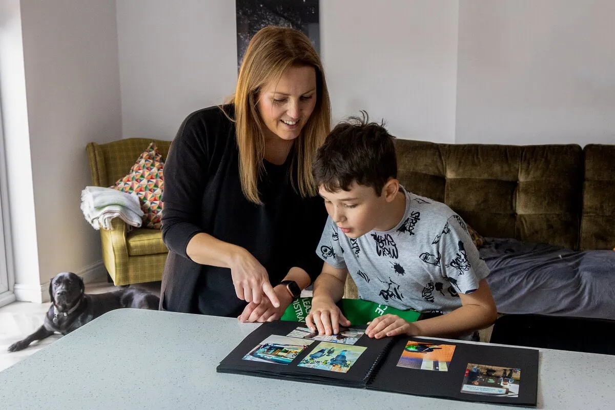 Parent and children at home looking through a picture book while a black labrador autism assistance dog rests behind them on the floor
